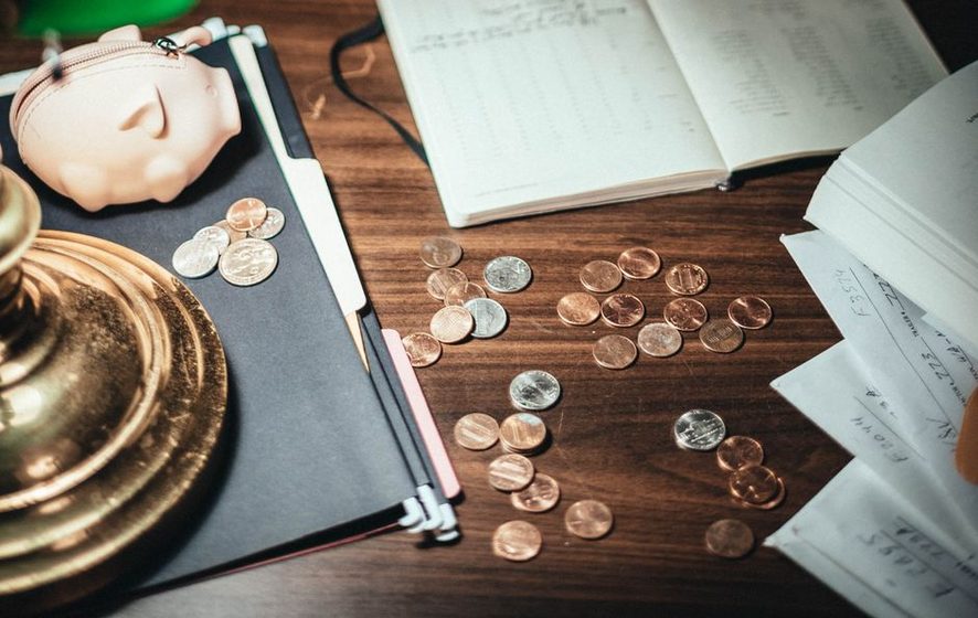 Mesa de madeira com um cofrinho de porquinho, um caderno de contas aberto e moedas espalhadas ao lado de recibos