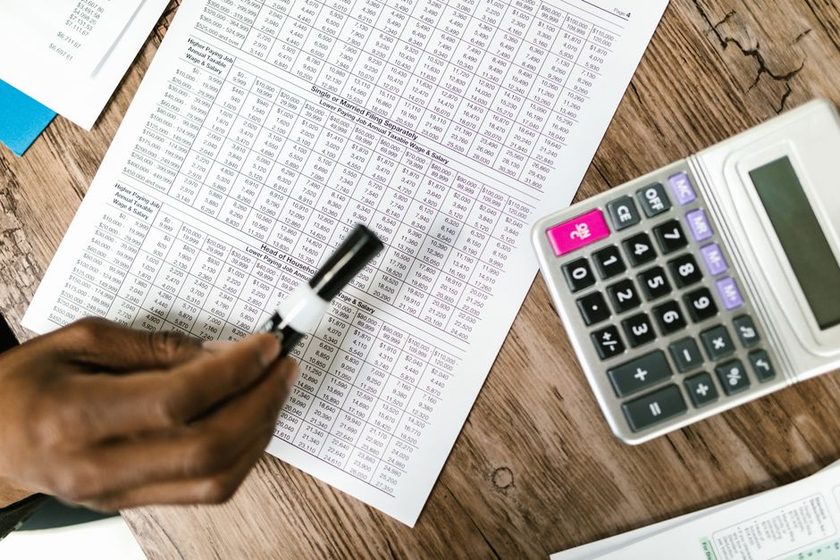 Mão segurando uma lupa sobre uma planilha impressa de orçamento ao lado de uma calculadora sobre a mesa de madeira