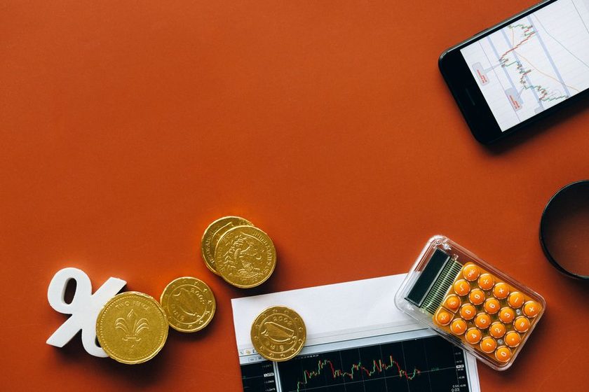 Moedas, um símbolo de percentual e um celular com gráfico de cotação sobre uma superfície laranja